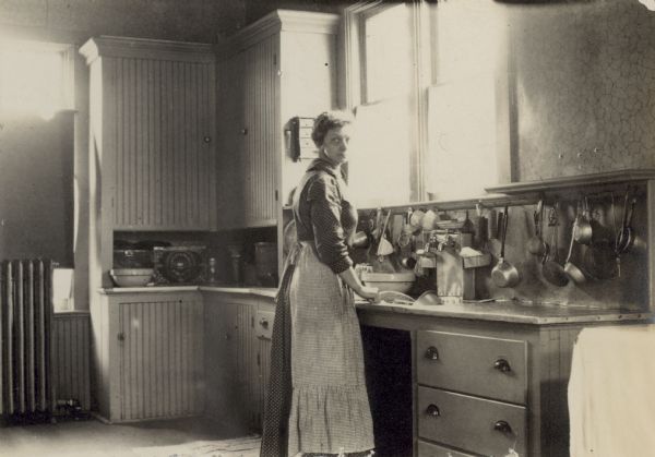Looking at the camera, Minnie Vent is standing in the kitchen in front of the sink. Sunlight is streaming in from the window above the sink. Kitchen cabinets and drawers are visible. A large tin of Huntley and Palmers biscuits is sitting on the counter. A radiator is standing on the floor in front of a window, which is covered by a shade. Kitchen utensils are hanging from nails above the counter.