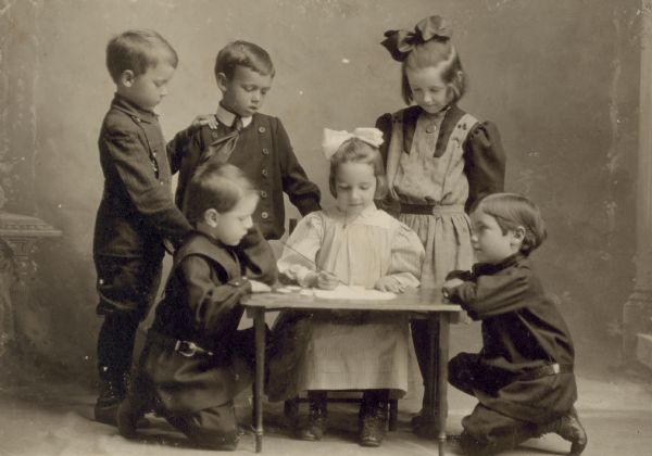 Group portrait in front of a painted backdrop of Eleanor Holt sitting at a small table,  writing or drawing on paper, with classmates kneeling and standing around her watching. Names (from left to right) Blair McQueen, William Galagher (kneeling), John Gill, Eleanor Holt (sitting), Elizabeth Galagher, Robert Harvey (kneeling). Caption reads: "Miss Emily McDonald, Teacher."
