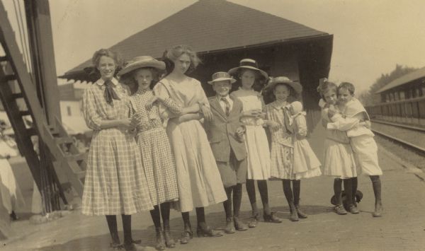 Eight cousins from the Holt and Stroh families are standing arm-in-arm at the depot. Caption reads: "Eight Cousins." Names from left to right: Margaret Stroh, Jeannette Holt, Harriet Stroh, Alfred Holt, Elizabeth Stroh, Juliet Stroh, Eleanor Holt, and Donald Holt.