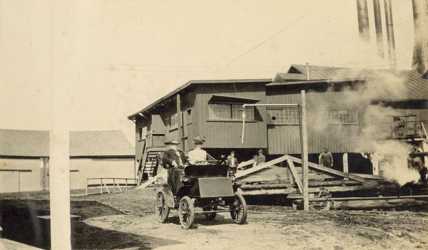 Lucy Rumsey Holt driving a car. Her father Captain I.P. Rumsey, a Civil War veteran, is sitting on the passenger side. Men are standing in front of the Holt Lumber Mill in the background. Caption reads: "Father and LRH at the mill."