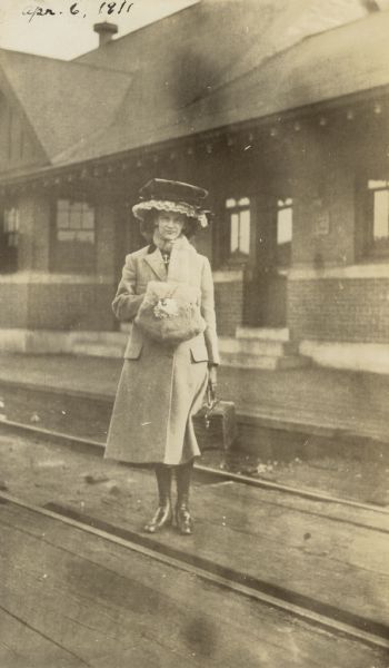 Dressed in traveling clothes, Lucy Rumsey Holt is standing by the railroad tracks at the Lakewood Station. Caption reads: "Just Starting for Lake Forest."