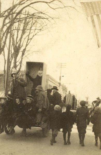 The Armistice Day parade is heading down Main Street. This float shows German Kaiser Wilhelm II's coffin. A group of joyful men and woman are riding on the back of the car. A line of boys are walking along with the float. Page caption reads: "Armistice Day - Main Street." Caption reads: "The Kaiser's Coffin."