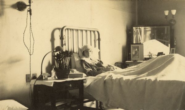 Eleanor Holt is lying in her hospital bed at Henrotin Memorial Hospital. Her left leg, under the bed sheet, appears to be elevated. There are several framed photographs on the bureau, including one of older brother Alfred Holt, who served in World War I, wearing his military uniform. The photographer's image is seen in the mirror, which is hanging above the bureau. Caption reads: "Mary Eleanor Holt."