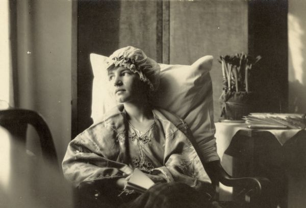 A portrait of Eleanor Holt with her face bathed in natural light, sitting in her hospital room. A pillow case is covering the back of the wooden chair. Wearing a silk jacket with frog closures and a night cap, she is looking out the window, with a book on her lap. A wool blanket is covering her lower body. There is a plant beside her on the table.