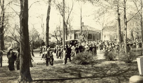 View across lawn towards the parade down Main Street, celebrating the return of the 32nd Infantry Division, Company M after WWI. A car is in the parade, followed by a marching band. A big crowd is welcoming the soldiers home. Page caption reads: "Return of Company M 32nd Red Arrow Boys - May 19 or 16, 1919."