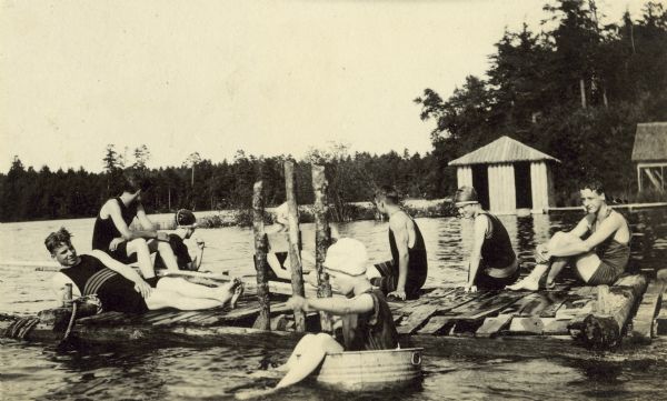 A group of eight swimmers are hanging out together at the Holt pier on Archibald Lake. A girl in the foreground is floating in a wash tub. The young men are wearing one-piece bathing suits covering their bodies from hips to shoulders. All of the young women are wearing bathing suits and tightly fitting swim caps. One young man is sitting on the wooden diving board. The bath house is across the water in the background.