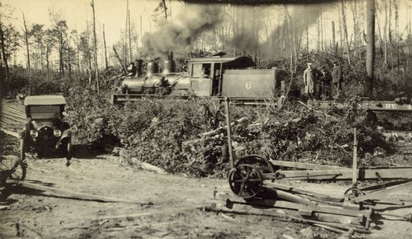 View of a locomotive on the logging railroad spur, with the engineer. Three men are standing on the tracks. An automobile is parked below on the left in front of some logs.