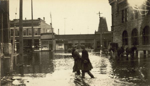 Two women are walking through floodwaters from the Oconto River. A horse-drawn carriage with its wheels halfway underwater is in the background near the Oconto County Courthouse and several commercial buildings.