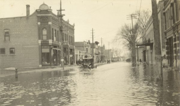 The car is plowing through flood waters on Main Street. Commercial buildings and telephone poles are in the background.