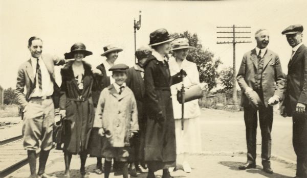 A group of Holt family members and others are standing by the train tracks. Alfred and Madeline Holt are standing on the far left. W.A. Holt is standing on the far right. The woman in the center holding the umbrella may be Lucy Rumsey Holt.