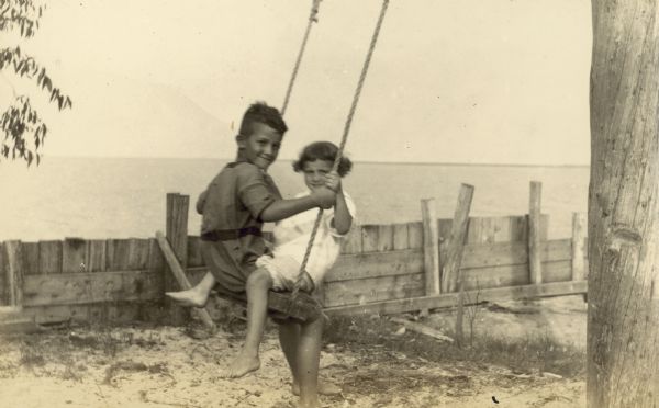 A boy and girl are swinging together with a lake in background. Page description reads: "Miss McDonald's Shack."