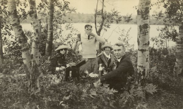 W.A. Holt and three of his adult children are picnicking under birch trees on the shore of Archibald Lake.

From left to right: Jeannette Holt, wearing fashion sunglasses, is sitting on the ground with the picnic box in front of her. Donald Holt, on his knees, is holding up a metal bottle and drinking from a cup. Eleanor Holt is holding a sandwich. W.A. Holt is holding a large metal bottle.