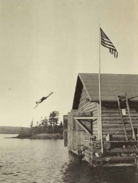 The person in midair, possibly David Rumsey, is diving from a height of 21 feet into Lake Michigamme. The log boathouse and American flag are in the foreground. Caption reads: "David 21 Feet."