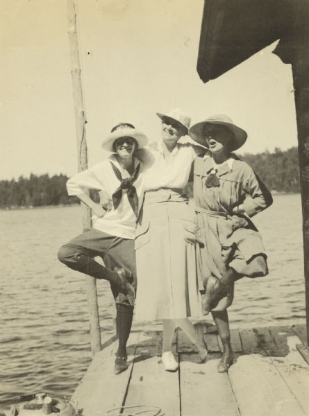 Three women are striking a theatrical pose on the pier. They are probably members of the Holt, Rumsey, and/or Stroh families. The wooden flagpole is on the left. The log boathouse is on the right. Lake Michigamme is in the background.