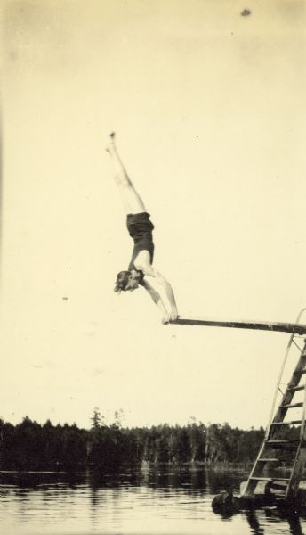 The diver, possibly David Rumsey, is doing a handstand on the end of the diving board. Archibald Lake is in the background.