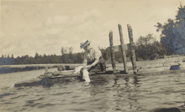 View from the lake of Miss Ransom washing clothes by hand, using lake water. Beside her on the pier is an enamel metal wash bowl. Archibald Lake is in the background. Caption reads: "Miss Ransom - Expert Laundress - Island Lodge Pastimes."