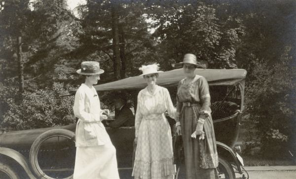 Sisters Lucy Rumsey Holt (l) and Julia Rumsey Stroh (c) with Maud Kimball are standing by a car at the I.P. and Mary Mathilda Rumsey family home. Behind them, a male driver is sitting in the car.