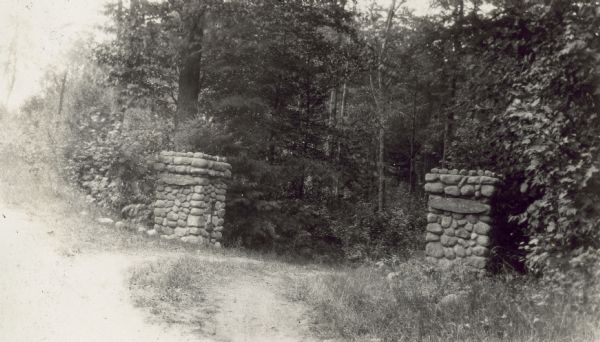 View of the Island Lodge stone gates. An "Island Lodge" sign is hanging on the left gate, and "The Ark" sign is hanging on the right gate. The long driveway leads down to the Archibald Lake shoreline and a pier where family and visitors would board a small boat and travel the short distance to the Island. Caption reads: "Stone Gates."