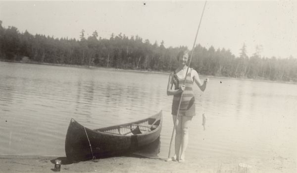 A woman standing on a shoreline is showing the fish she caught on the hook with the cane pole. A wooden canoe is beached beside her on the sand.