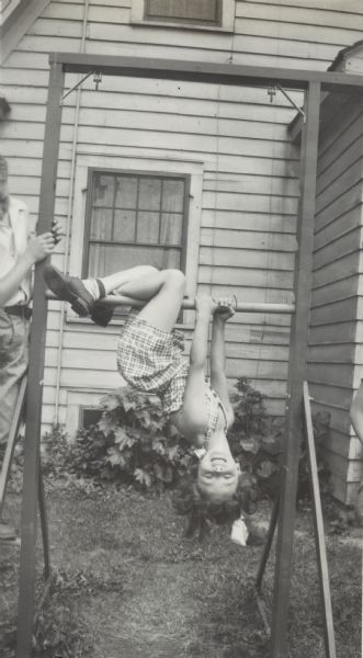Eunice Blanchard is hanging from the horizontal bar in the yard. A man, (partially out of frame) possibly her father, is watching her play.