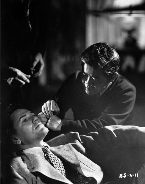 Cinematographer James Wong Howe applying makeup to actor John Garfield who is sitting in a chair on the set of the 1947 film "Body and Soul."