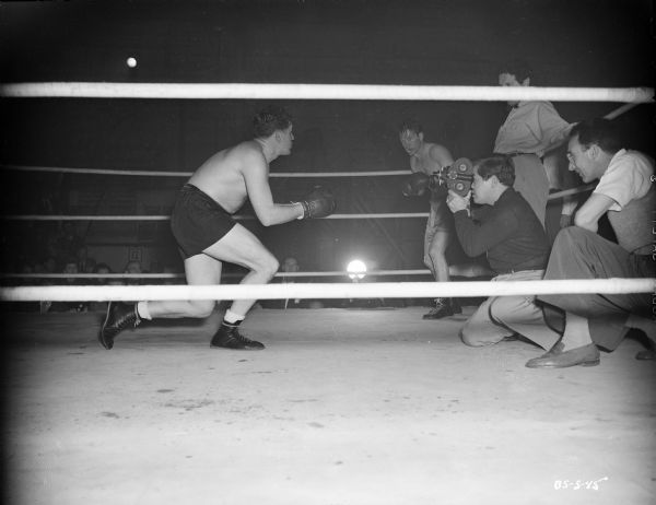 James Wong Howe filming John Garfield with a handheld camera in a boxing ring during the making of the 1947 film "Body and Soul." Two actors are standing behind Howe, and another man is kneeling next to him.