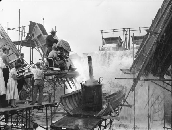 View of people filming a scene from the 1951 film "The African Queen." A partially built boat is being splashed with vast amounts of water. The camera crew is watching from the left.  