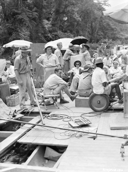 The crew resting on the set of the 1951 film "The African Queen." Katharine Hepburn, holding an umbrella, is standing and talking to someone in the center of the group.