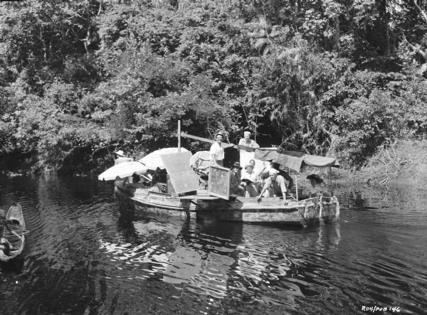 Cast and crew of the 1951 film "The African Queen" on a boat, preparing to film a scene. They are floating on a body of water, with trees behind them. Actors Humphrey Bogart and Katharine Hepburn are under a cloth cover on the right side of the boat.