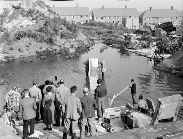 Assorted members of a film crew by a lake, during the making of the 1951 film "The African Queen." Two men are standing in the center of the image, holding what is probably a large mirror. A movie camera is on the left of the image, almost hidden from view by a few people.