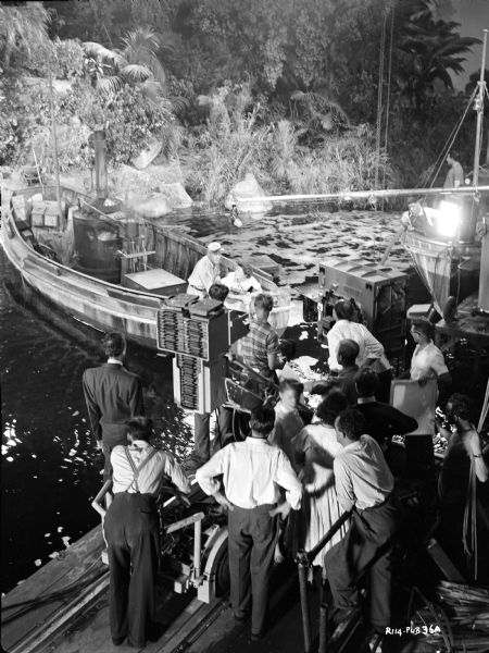 Crew, in foreground, filming a scene for the 1951 film "The African Queen." Humphrey Bogart and Katharine Hepburn are in a boat with a microphone over their heads. The boat is on a body of water, and the crew are standing on what is probably a dock. 
