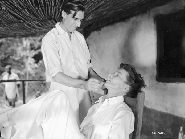 Katharine Hepburn having her makeup applied by makeup artist George Frost while on set of the 1951 film "The African Queen." They are on a covered porch. 