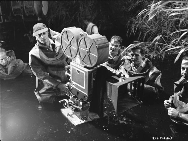 Film crew for the 1951 film "The African Queen," including John Huston, Ted Moore, and Edward Scaife, surrounding a movie camera. They are all partially submerged in water and are wearing diving suits.