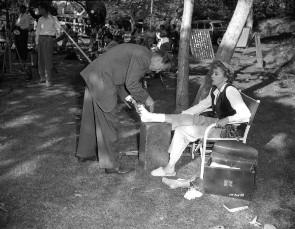 Director Ida Lupino sitting in a chair, getting her foot bandaged by an unknown man on the outdoors set of the 1949 film "Never Fear." A foot cast is next to her. Film equipment and some men, probably the film crew, are in the background.