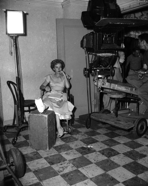 Director Ida Lupino is sitting in a wheelchair on the set of the 1949 film "Never Fear" with her right foot in a cast; her foot is propped up on a block. A movie camera is on the right side of the image.