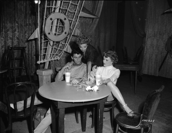 Director Ida Lupino is standing behind actors Keefe Brasselle and Sally Forest. Brasselle and Forest are sitting at a table with Lupino bending over them. They are on a nautical-themed nightclub set during the making of the 1949 film "Never Fear." 