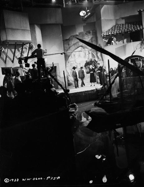 Cast and crew are filming a scene from the 1938 movie "Blockade." A microphone is hanging over the cast in the background. In the left-foreground is the crew. The set resembles a harbor.