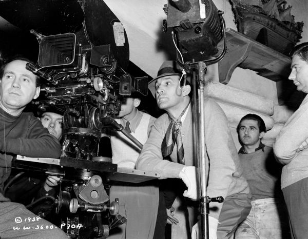 Director William Dieterle in between a movie camera and light with some of the unidentified film crew on the set of the 1938 film "Blockade." A model ship is above his head in the upper right corner.