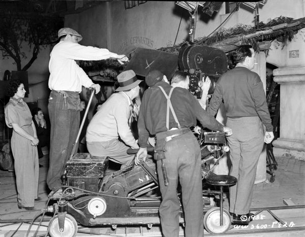 Director William Dieterle is sitting on a camera dolly on tracks with unidentified crew. They are filming a scene from the 1938 film "Blockade."