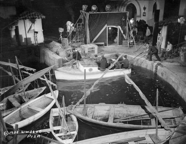 Cast and crew preparing to film a scene from the 1938 film "Blockade" on a sound stage. The set is made to look like a dock, with a group of actors in a boat floating in a tank of water. The set is surrounded by lights. 