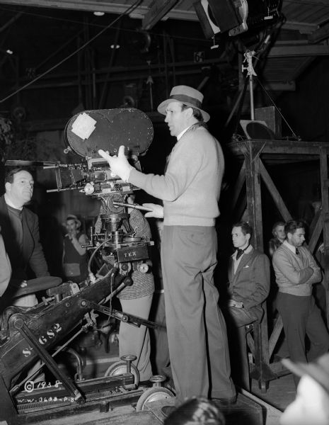 Director William Dieterle is standing next to a camera dolly, with his hand on a lever attached to the movie camera. He is surrounded by a film crew on a sound stage, during the production of the 1938 film "Blockade." 