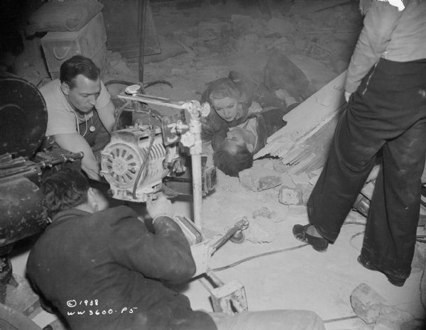 Actors Henry Fonda and Madeleine Carroll are lying on the ground surrounded by rubble. They are preparing to film a scene from the 1938 film "Blockade." Three unidentified men, part of the film crew, are setting up a movie camera and light at ground level.