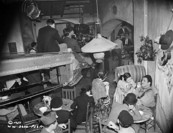 The crew for the 1938 film "Blockade" are filming a scene in a cafe set. The movie camera is on a crane over a crowd of extras.