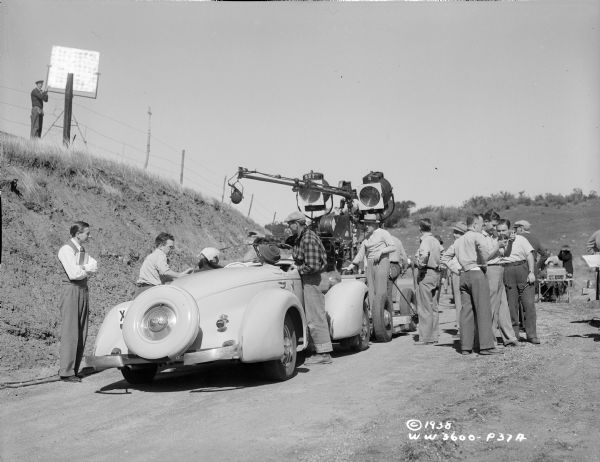 Cast and crew are filming an outdoors scene from the 1938 film "Blockade." Two actors (probably Madeleine Carroll and Henry Fonda) are sitting in a car, while lights, a microphone and a movie camera are set up. Director William Dieterle is standing nearby. There is a man standing next to a reflector on a hill on the left.