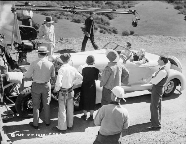Elevated view of director William Dieterle standing and instructing actors Henry Fonda and Madeleine Carroll. The latter two are sitting in a car facing a movie camera, while a microphone is hanging over them. They are in an outdoor scene for the 1938 film "Blockade."