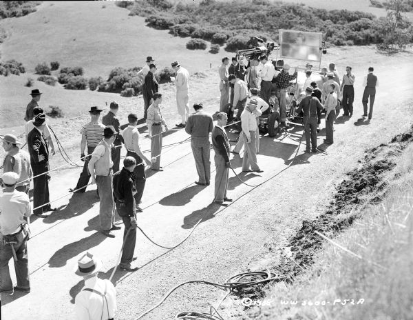 Henry Fonda and Madeleine Carroll are sitting in a car in the background, and director William Dieterle is standing in the center. They are shooting a scene outdoors for the 1939 film "Blockade." The film crew is holding up cables that are presumably powering the film camera and microphone, both of which are in front of the car.