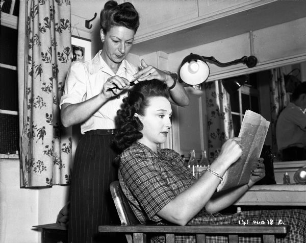 Actress Googie Withers is having her hair done by an unidentified woman, probably hair stylist Ivy Emmerton, on the set of the 1948 film "Miranda." They are in a dressing room. Withers is sitting in a chair reading a newspaper.