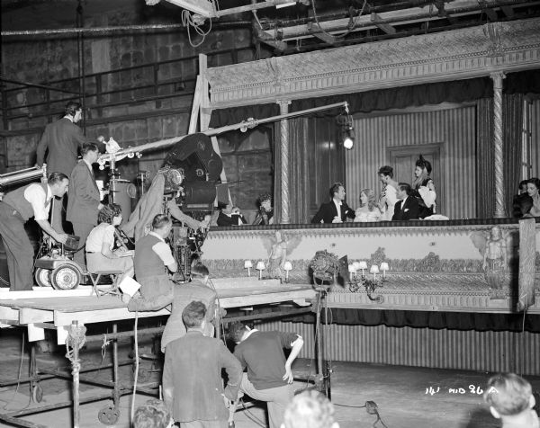 Cast and crew preparing to film a scene on a theater auditorium set for the 1948 film "Miranda." Actors Griffith Jones, Glynis Johns, Googie Withers, John McCallum, and Sonia Holm are sitting in a box. On the left side is an elevated platform, and on top of it is a movie camera, a microphone, and some of the film crew.