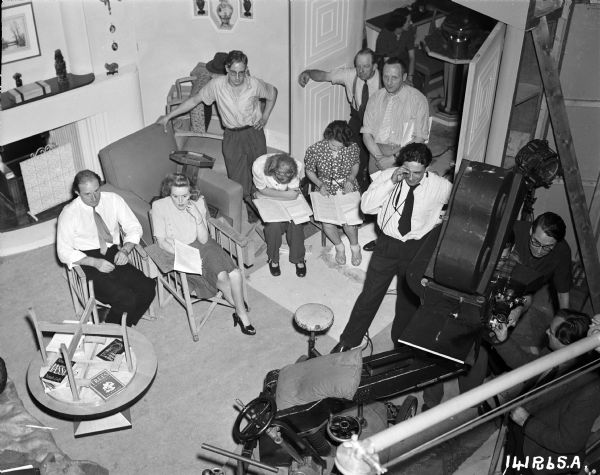 Elevated view of the crew watching the making of a scene from the 1948 film "Miranda." The scene itself is not shown. Producer Betty Box is second from the left. The set's framework can be seen on the far right, next to the movie camera and a crane.