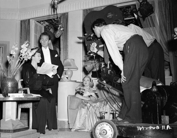 Actors Googie Withers, Griffith Jones and Glynis Johns filming a scene from the 1948 film "Miranda." Withers is sitting in a chair with a newspaper, Jones us standing next to her, and Johns is reclining on a couch. A microphone on a boom is suspended above Withers and Jones. On the right are men standing behind a movie camera  on a dolly.
